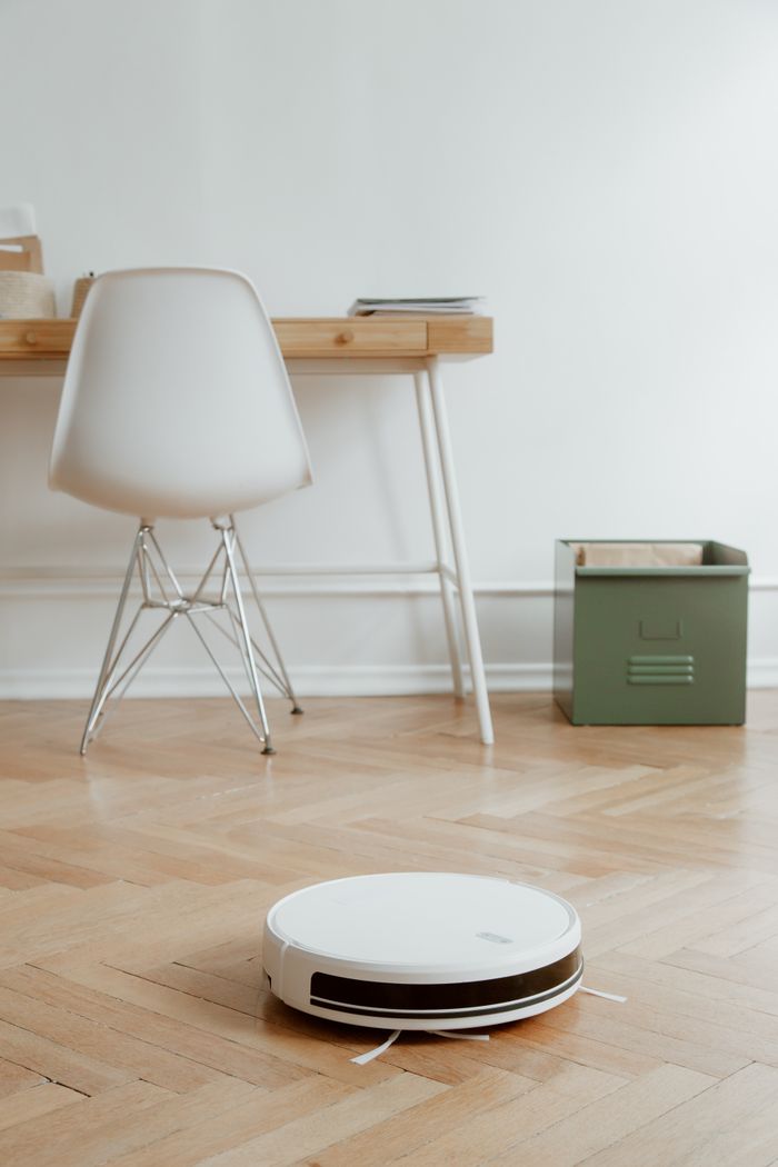 services-01 A modern minimalist room featuring a robotic vacuum cleaner on a wooden floor beside a desk and chair.