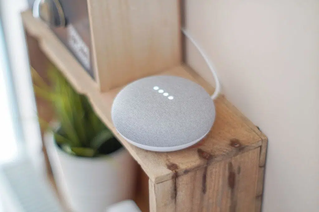 about-us-02 A Google Home Mini smart speaker on a wooden shelf, blending technology with home decor.