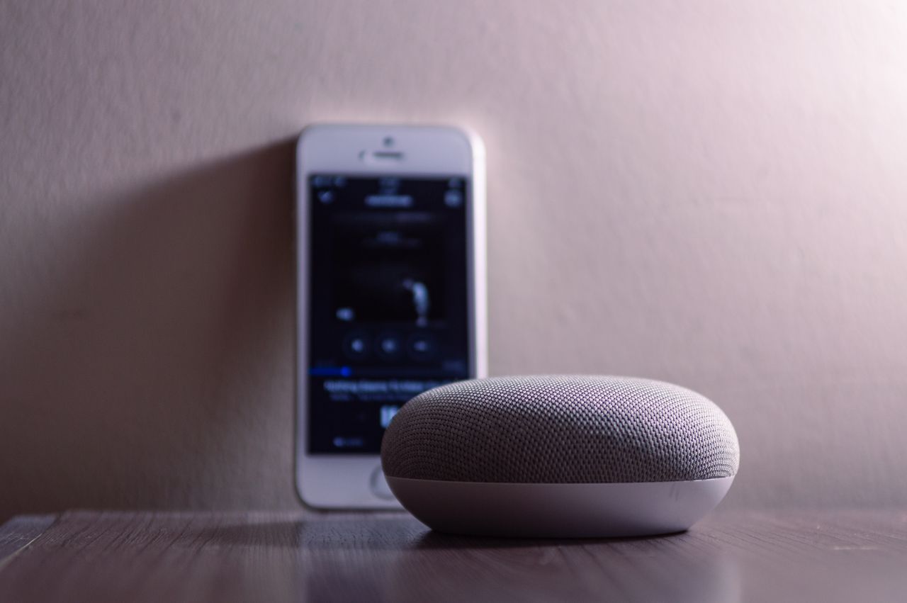 heros-img A modern smart speaker and smartphone on a wooden table, indoors.