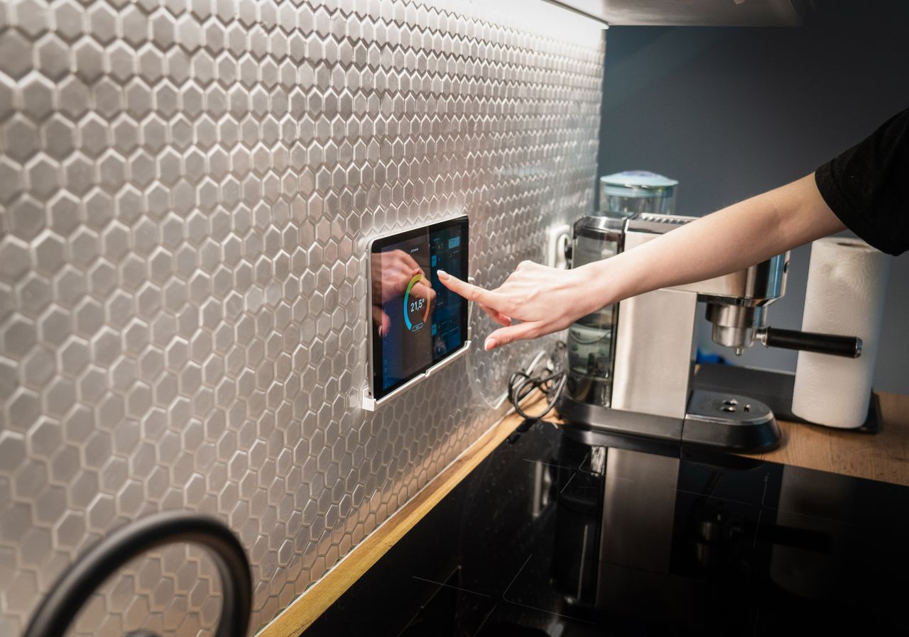 about-us-01 A modern kitchen with a touchscreen smart home control panel integrated into a hexagon-patterned wall.