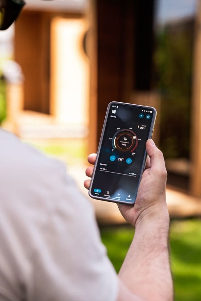 A man uses a smartphone app to control a modern sauna outdoors, showcasing sauna tech innovation.