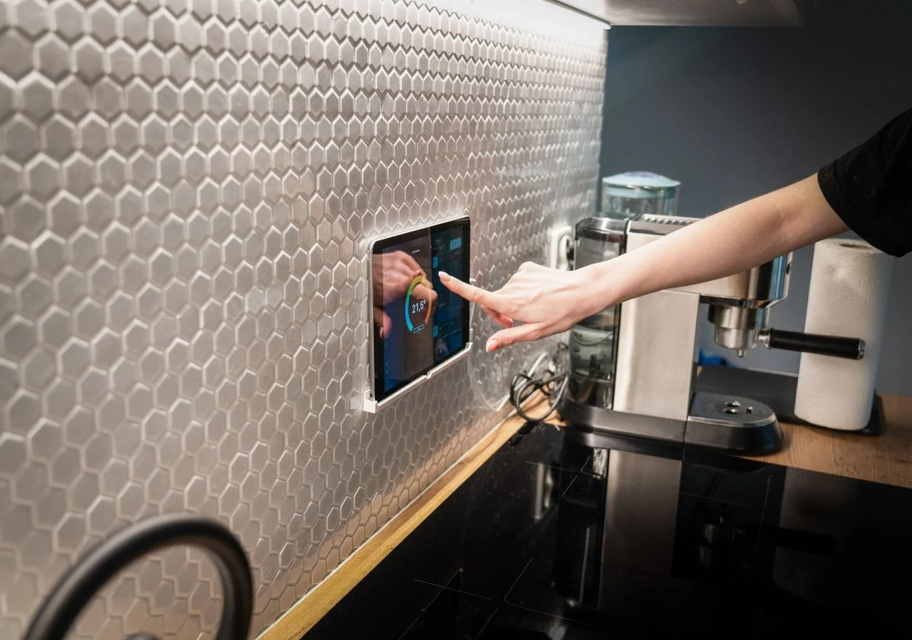 heros-img A modern kitchen with a touchscreen smart home control panel integrated into a hexagon-patterned wall.